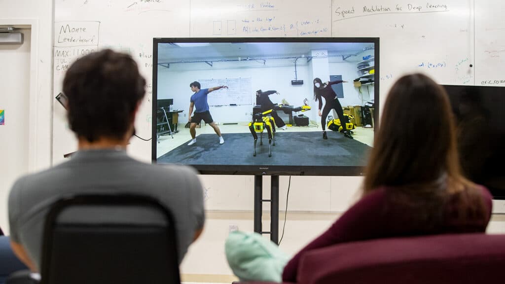 Several people facing away from camera watch a monitor showing three students dancing with Spot