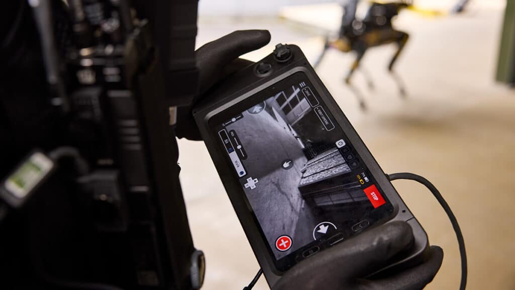 A close up on an operator's hands holding a tablet controller showing the view from a robot in frame behind