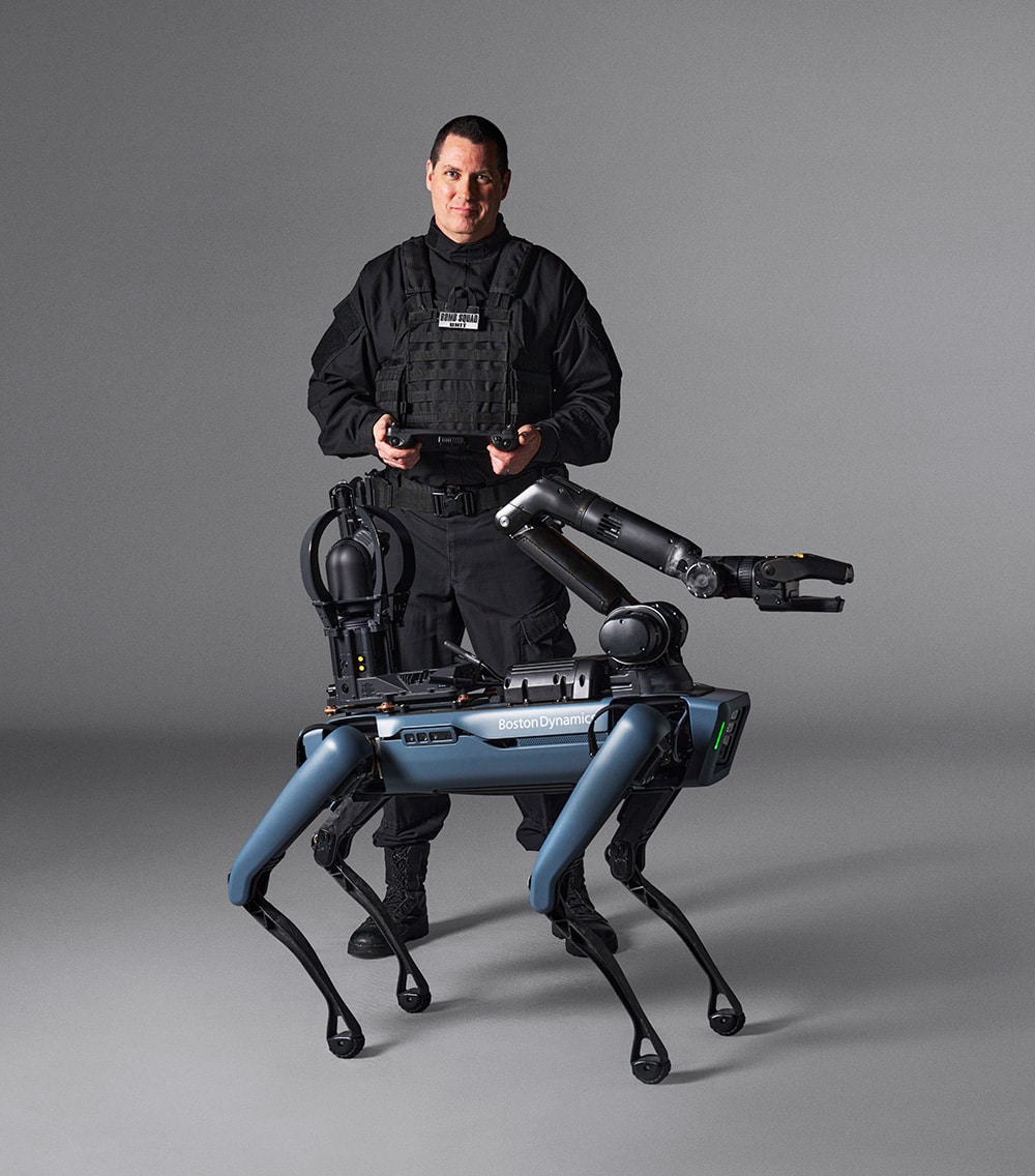 A police officer holding the tablet controller stands behind a blue Spot robot with an arm and camera payload