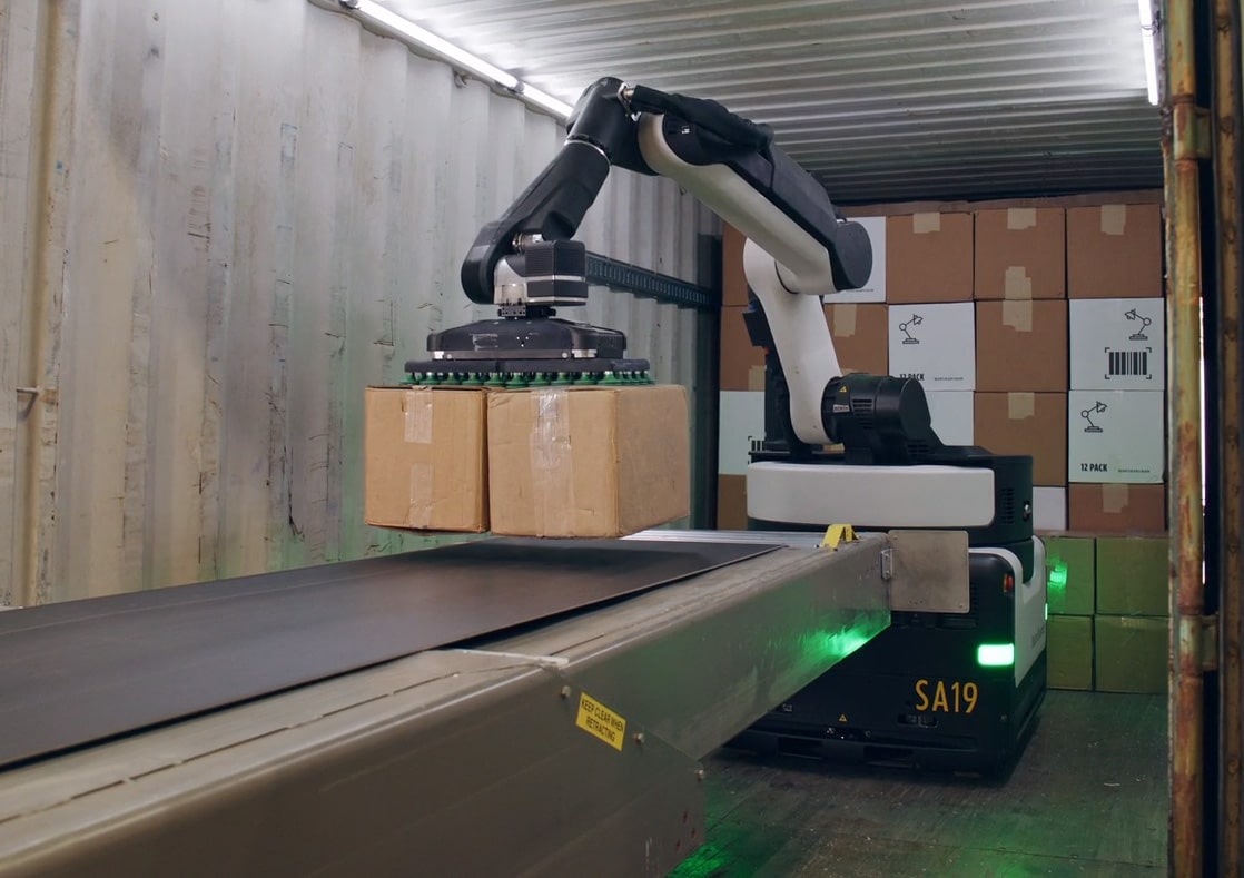 Stretch robot grasping two brown boxes simultaneously with the suction gripper at the end of an industrial arm, hovering over a conveyor belt inside a container