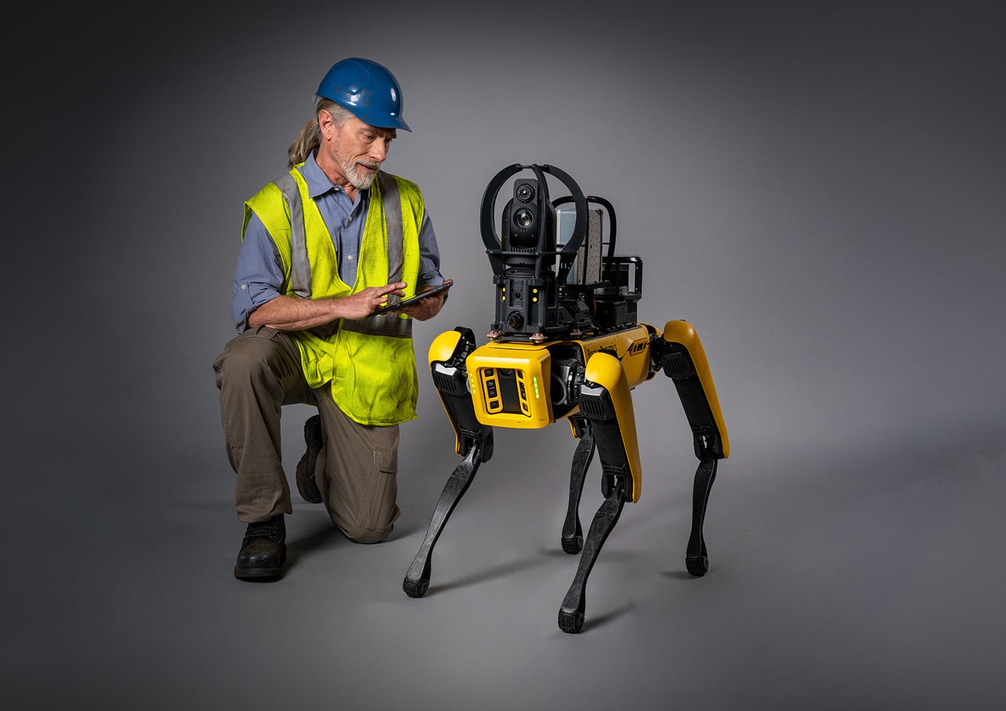 A worker kneels next to Spot, adjusting setting on the tablet controller